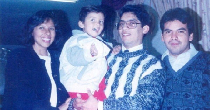 A slightly faded photo of a Latino man with glasses, smiling. holding a little boy with a jacket, red pants and brown hair. He is flanked by a smiling Latina with a bob haircut and a black jacket, and another man with dark hair and a faint mustache. 