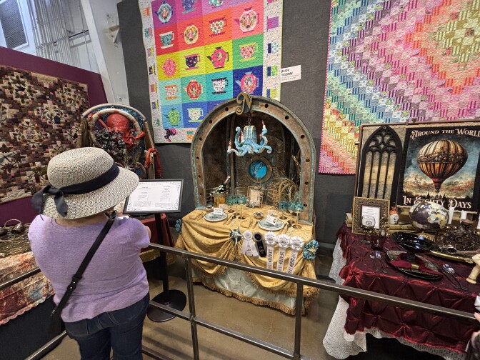 A woman in a purple t-shirt and gray straw bucket hat with black bow looks at table setting displays. In the center is an ocean-themed table with a blue octopus chandelier, gold draped tablecloth, and a porthole in the background.