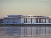 A large, low white building sits on the bank of a river. It is pictured from across the river.