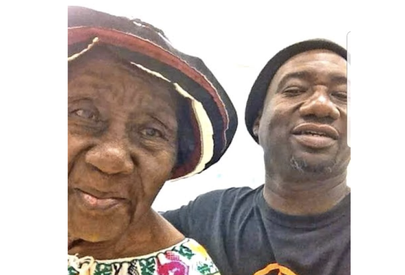 A Black man and a Black woman smile for the camera in a selfie. The man is on the right, and has a salt and pepper beard, he's wearing a black beanie. The woman is older, she has a striped red, white and black hat on, and a floral white dress with pink, green and yellow designs.