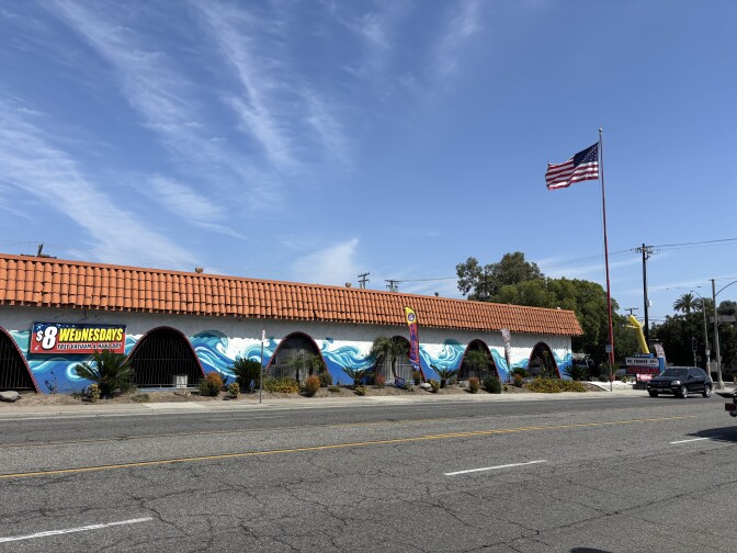 A car wash with a Spanish tile roof and white walls with cut out arches and waves painted round the arches. An American flag on a pole is at the corner of the building. 