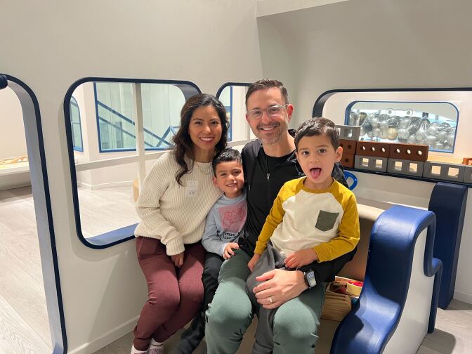 A family of four, including a female wearing red pants and a cream sweater, a young male in a blue and red t-shirt, a male wearing glasses and a black hoodie and a young male wearing a yellow, khaki and cream tshirt sticking his tongue out, smile at the camera. 