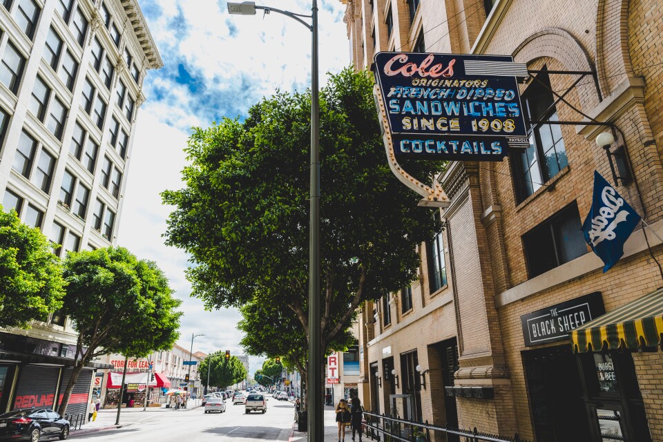 A sun-drenched downtown L.A. street scene with the iconic vintage neon sign of Cole’s — "Originators of the French Dipped Sandwiches Since 1908" — glowing proudly above the sidewalk. 