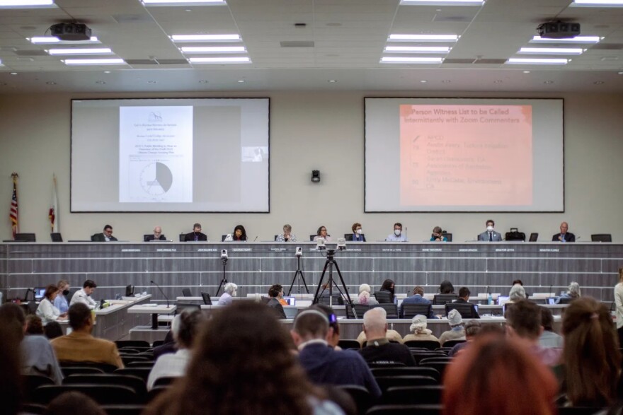 A row of people sit at a long dais, behind them are two large screens. Many people are pictured from behind, facing the dais.