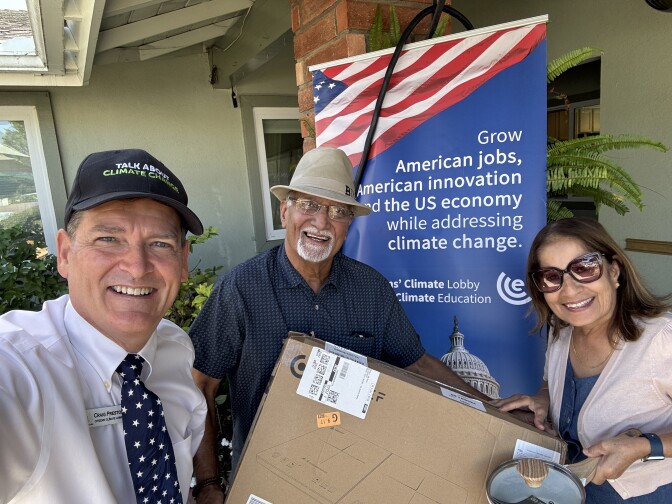 A white man wearing a black baseball cap reading "talk about climate change" and a white shirt and blue tie with white stars smiles taking a selfie with a man and woman who hold a cardboard box with a drawing of an induction cook top. A blue sign with an American flag in the background reads "Grow American jobs, American innovation adn teh US economy while addressing climate change."