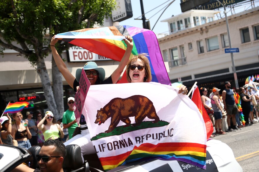 Two women sit on the back of a convertible. Each holds colorful flags.