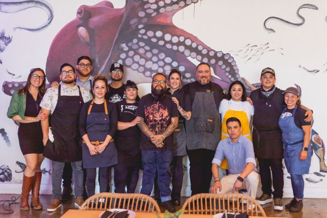 A group of people made up of men and women, stands up against a large mural of a purple octopus. All the people have various complexions of medium to dark skin, with some wearing aprons and others wearing more casual attire.  