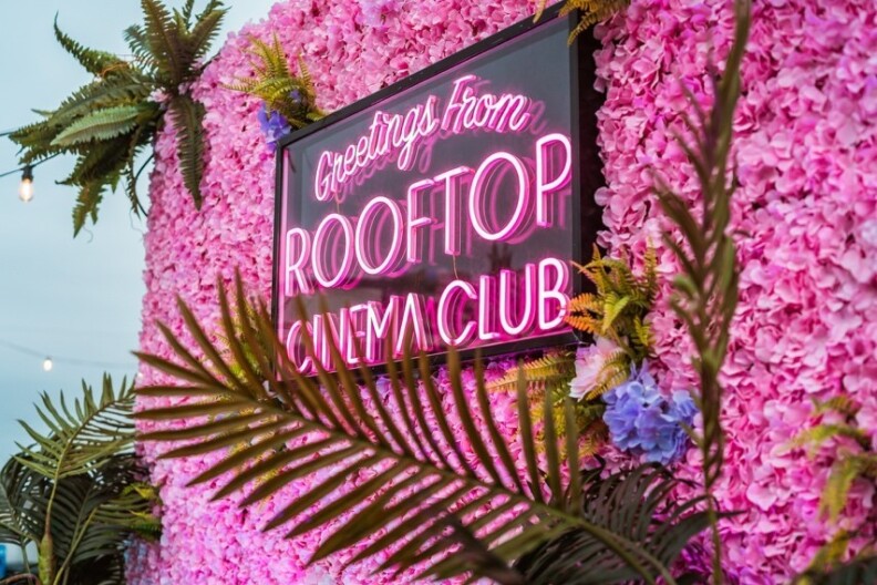 A neon sign reads, "Greetings from Rooftop Cinema Club, surrounded by a wall of pink flowers and palm branches.