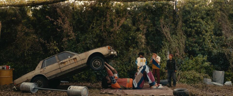 Photo of a large man holding up a car with three people staring at him.