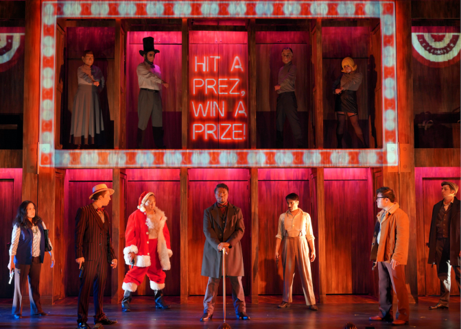 The cast of the musical "Assassins" are on a stage, several of whom are standing inside cubicles built into a two-level structure.