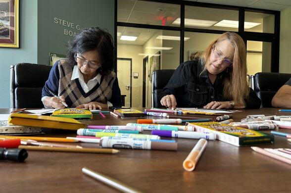 Two people color at a conference table covered with markers and colored pencils, near a wall reading 'Steve G... Board.'