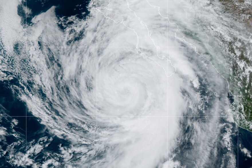 A satellite image shows the Mexican coastline in greens and browns overlaid with a giant white vortex of clouds centered just offshore over the ocean. Tendrils of the hurricane twist out over the land.