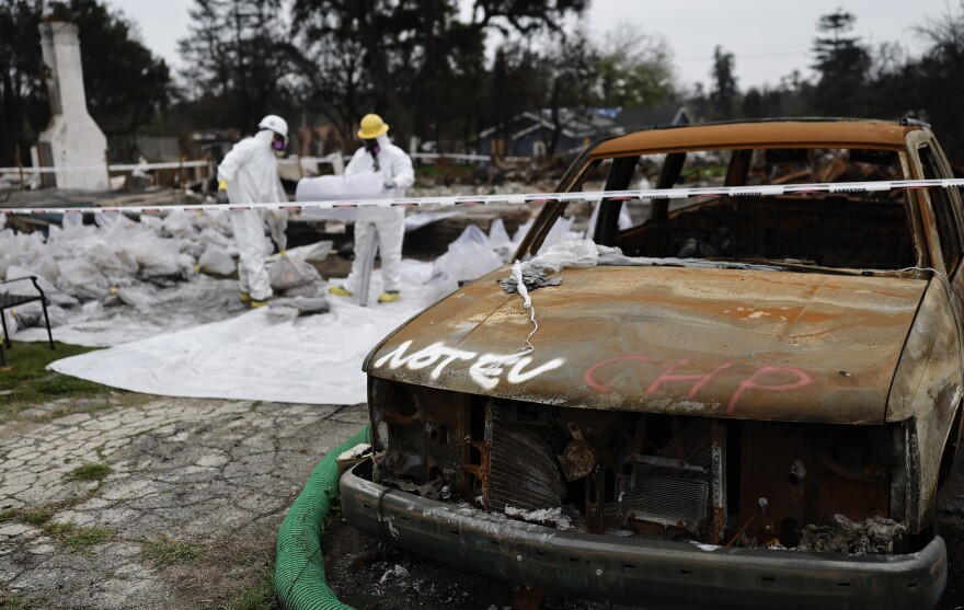 U.S. Army Corps of Engineers contractors in protective gear at a destroyed home in Altadena. They stand near a burned car marked "Not EV."