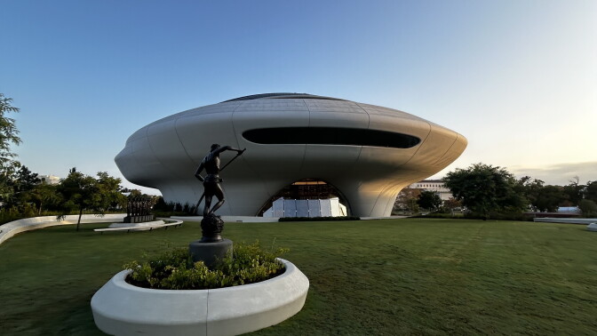 A saucer-shaped building sits on a lawn behind a bronze statue of a figure holding a pole atop a pedestal.