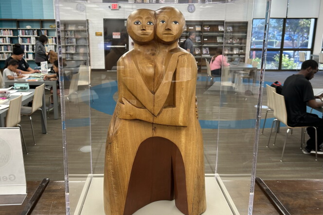 A wood sculpture of two people, they embrace and look at the viewer.