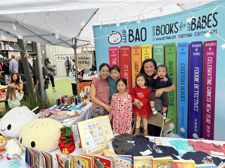 Two Asian American women and four children smile and stand in front of a display of books. There are two giant plush bao buns on the table. The sign behind them reads Bitty Bao. Bitty books for bitty babes. 