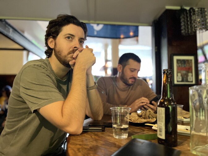 Two white men with goatees sit at a bar table looking toward a screen off-camera.