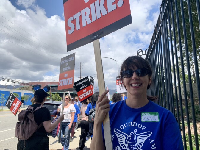 A Latina woman in a blue shirt that reads "Writers Guild of America" holds a sign in front of a fence while a group of people picket behind her. 