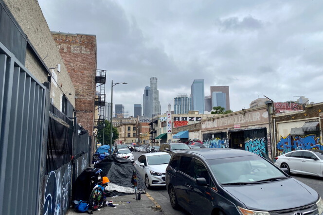 The LA skyline as seen from Skid Row. There are tents lining one side of the sidewalk next to parked cars.