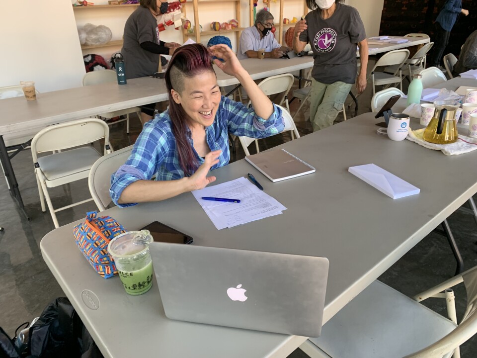 A person with long pink hair that's shaved on the side smiles while looking at a laptop open to a Zoom meeting. 