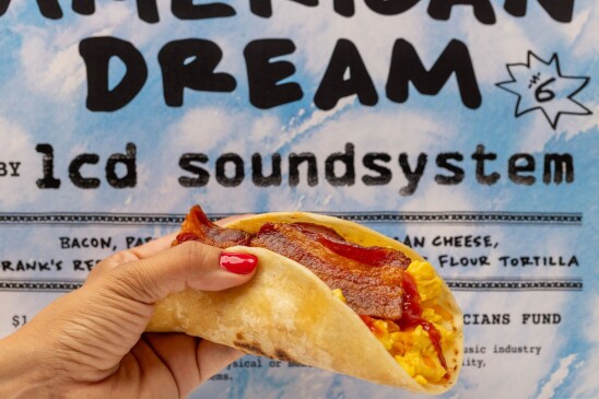 A female hand with red nail polish holds a taco with egg and bacon. In the background is a blue and white poster advertising the taco, including the "American Dream" and its price — $6.