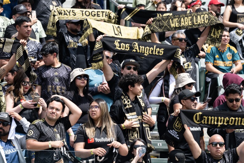 A close-up shot at LAFC fans in the stands from earlier this year , many wearing the team's black-and-gold colors in the form of bucket hats and jerseys and waving teams flags that say "Los Angeles," "We Are LAFC" and "Campeones"