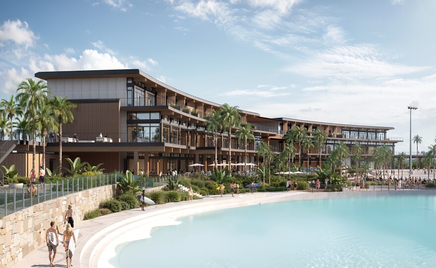 The exterior of two surf pools are lined with palm trees and building with patios where onlookers can catch the action.  People are walking along the edge of the pools, with some holding surfboards. 