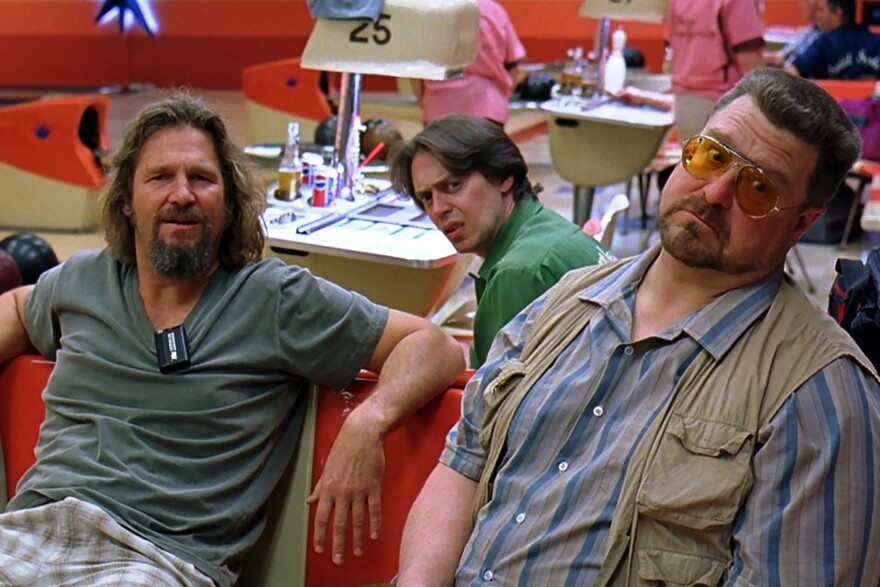 Three men sit in a bowling alley, looking toward camera in a still from the movie 'The Big Lebowski.' 
