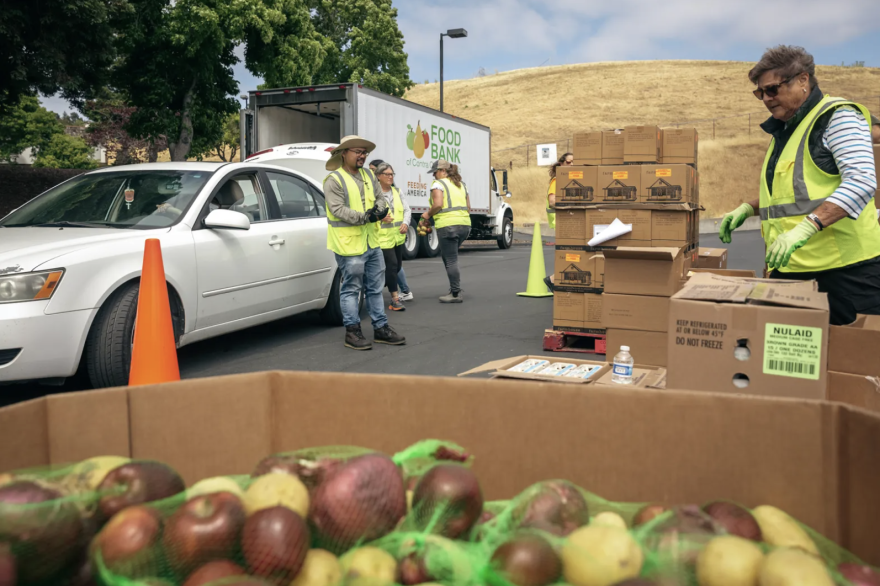 People in bright green jackets are outside and surrounded by boxes of food. A white car sits nearby with its trunk open.