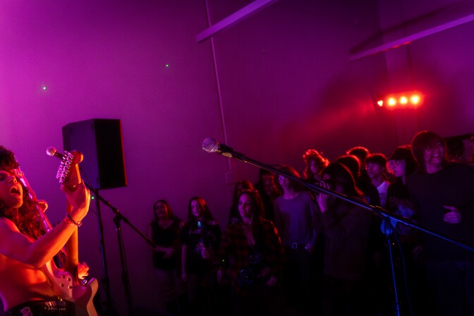 The band Saints of Sinners plays on stage under the glow of orange lights. The guitarist has long hair and is shirtless. 
