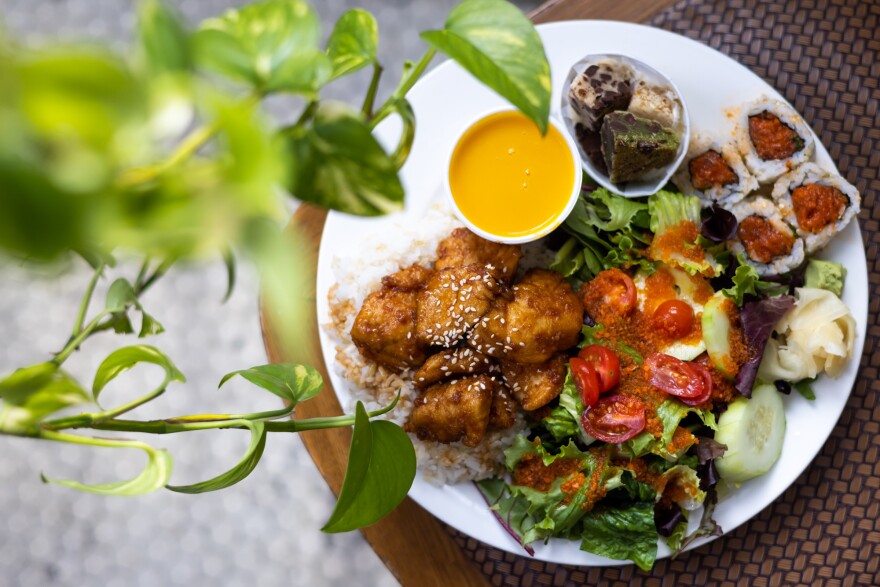 A white plate on a tabletop contains chicken over a bed of rice, a small cup of yellow soup, a paper cup of cookies, four sushi rolls, and a green salad with small sliced tomatoes covered in dressing. To the left is a green plant vine.