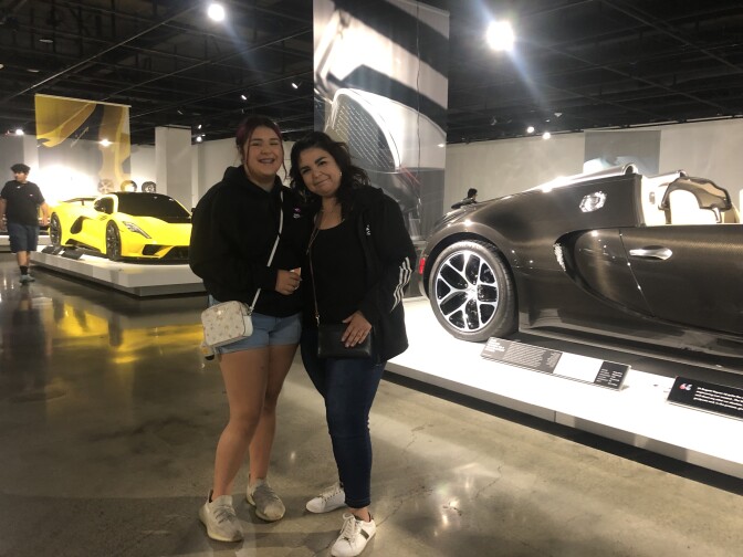 A young woman and her mother stand in a car museum. Fancy sports cars are behind them. 