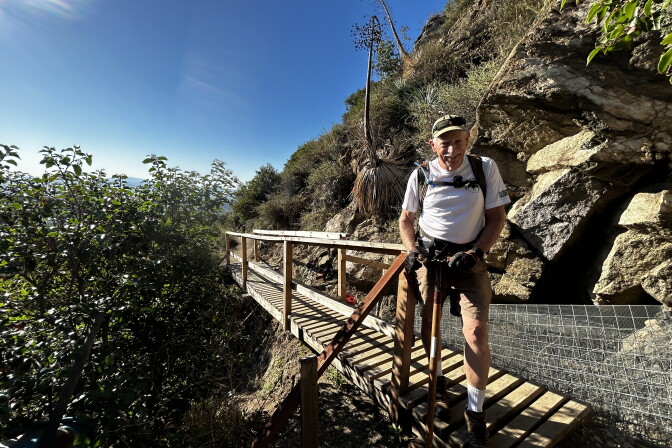 An older white man wearing a tan baseball cap and white t-shirt and tan shorts stands at the end of a new wood bridge smiling on a sunny day. In the background are large boulders, greenery and yucca plants. 