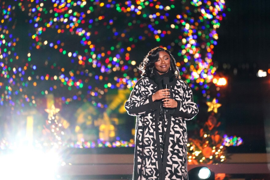 Samara Joy, dressed in long, warm a black-and-white patterned coat, performs in front of Christmas lights and trees.