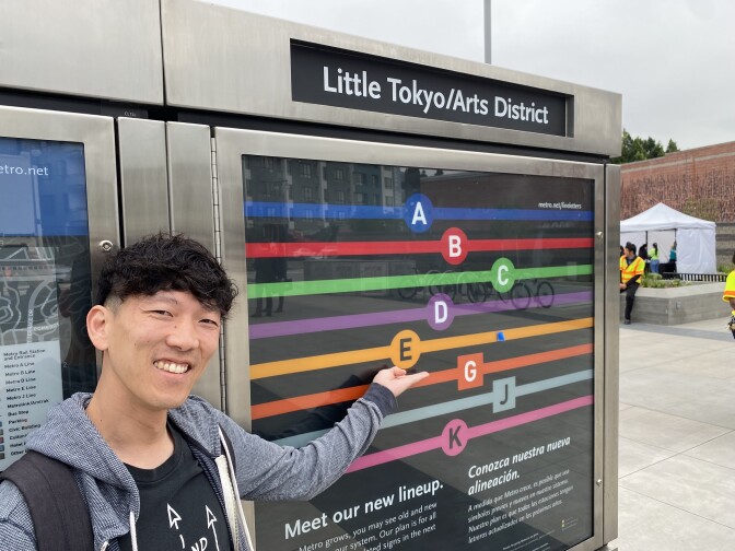 An Asian man wearing a grey sweatshirt over a dark shirt faces the camera while gesturing with his left hand to a sign for the new Little Tokyo/Arts District Metro station, which shows the several different color-coded lines that run through the station.