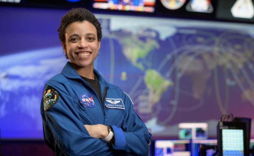 Jessica Watkins stands in a NASA blue jumpsuit, arms crossed, with a big picture of the earth behind her