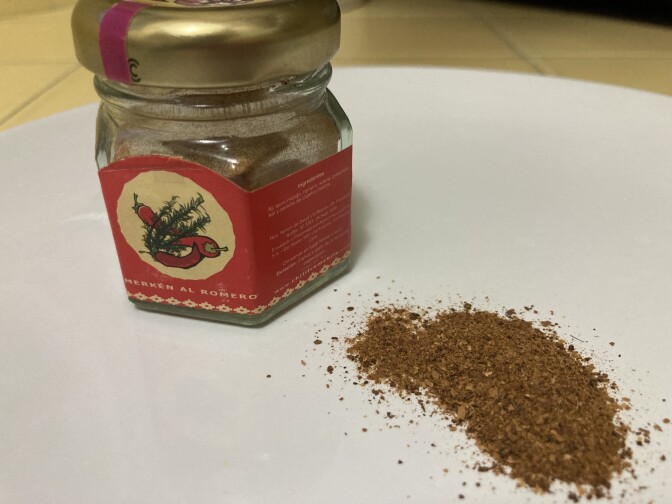 A glass jar with a bright red label showing red chili peppers sits next to a small pile of chili powder. Both are on a white plate