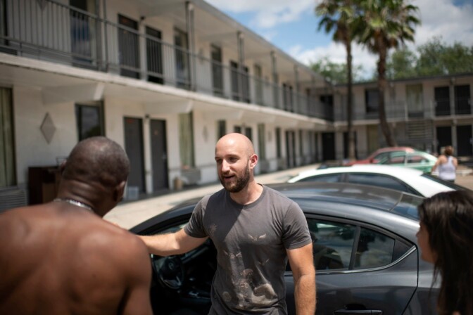 Casey Malich stands in the parking lot of a white motel with dark gray doors, propping his dark gray car door open as he speaking a shirtless individual.