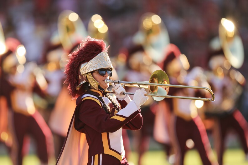 The USC marching band performs in their cardinal, gold and white uniforms. Most of the band is out of focus, with one trumpeter in focus at the center of the frame.