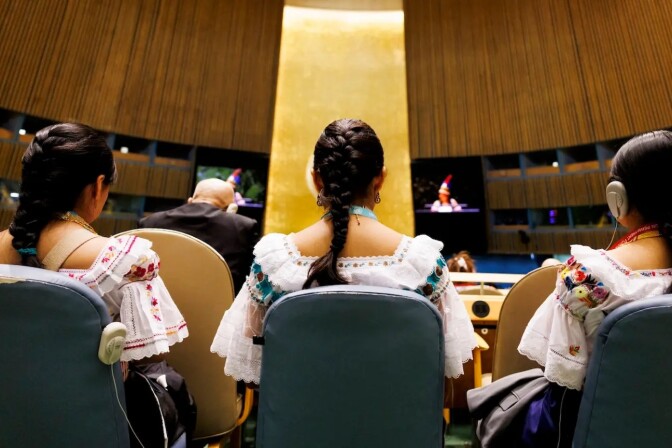 Three women are pictured from behind while sitting in blue chairs in a large, circular room. They each wear their hair in a single braid and are wearing white dresses with colorful embroidery. 
