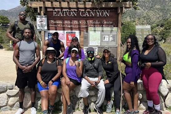 A group of Black women and men stand in front of the wooden sign for Eaton Canyon during the daytime, dressed for a hike. 