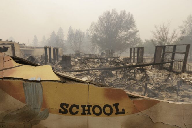 A burned down building with ashy air and burned trees in the background. The words "school" remain on a partially intact wall. 