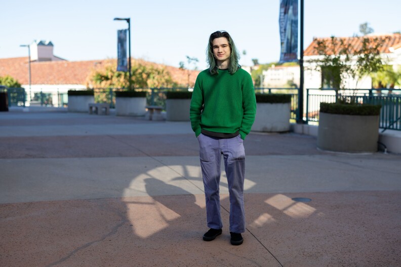 A masculine presenting person with shoulder length green hair wears a dark green sweater and purple pants while standing in a concrete college courtyard.