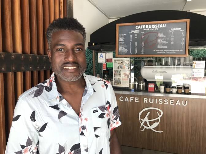 Photo shows a portrait of Edward Ackah-Miezah standing in front of the counter at Cafe Ruisseau, smiling and wearing a short-sleeve light-colored collared shirt with a flower motif.