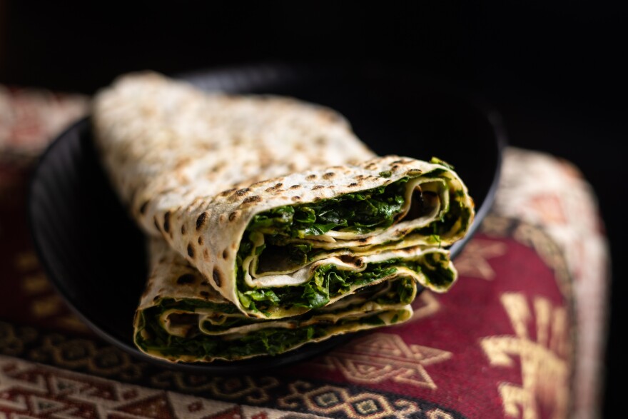 A light brown flatbread with tiny circular grill marks is stuffed with green herbs and vegetables and sits on a black-colored plate. The plate sits on a red tapestry with gold designs.