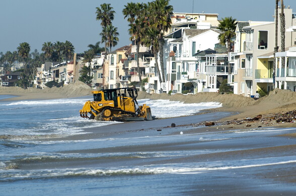 A tractor sits in the surf, with a sand berm and houses beyond it.