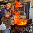 A woman in a brown Lomo Fuego apron stirs a wok over a powerful outdoor burner, producing dramatic flames that leap several feet into the air in a backyard restaurant's  patio area. 