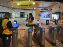 The interior of an underground public transit station. Two men in green and orange high-visibility vests and long pants are standing around the silver metal turnstile gates. The man on the left is working on a laptop computer at the same time. The station is largely empty and well-lit, except for one woman walking in the background. 