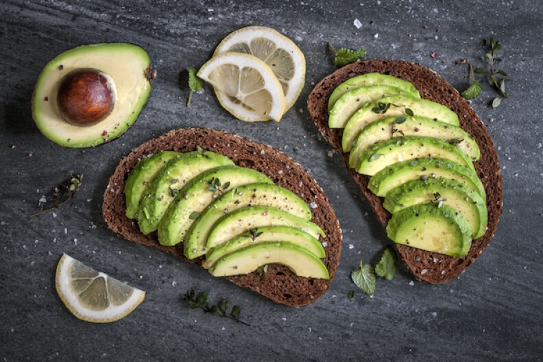 Rye toast with avocado and flaky salt displayed on a table. There are thing lemon slices and half an avocado with the pit still in pictured as well.