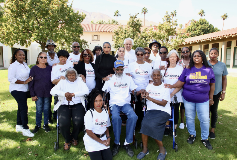 A group of people, many of them elderly, most wearing white t-shirts, pose for a photo on a grassy lawn.
